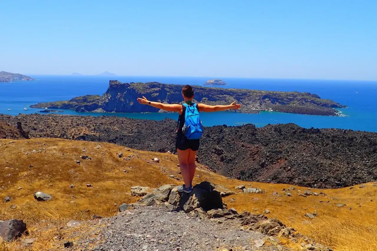 Widok na kalderę po trzęsieniu ziemi na Santorini. Kobieta z plecakiem stoi na wzgórzu, rozkładając ramiona.