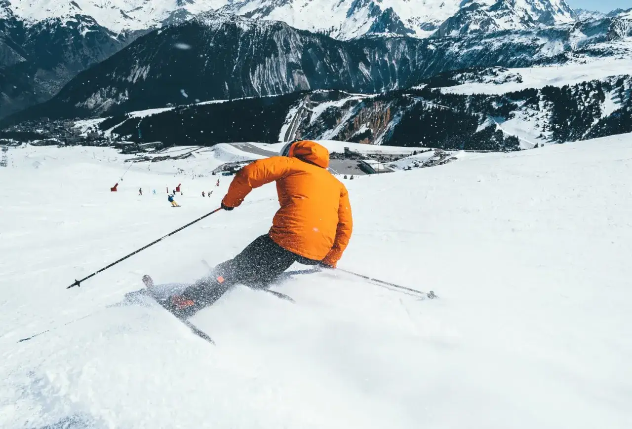 Narciarz w pomarańczowej kurtce zjeżdża po stoku, wzbijając śnieg. Widoczna jest tylna część narty i kijki.