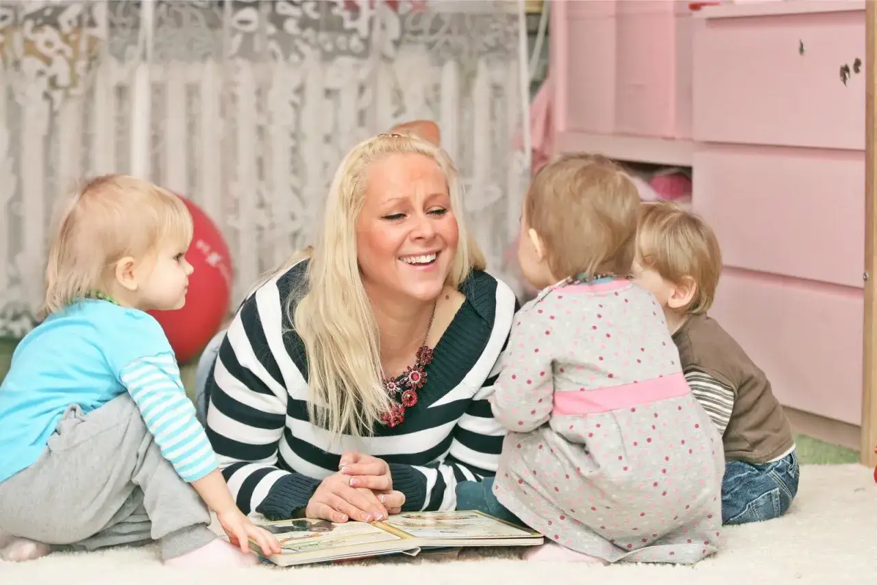 Uśmiechnięta opiekunka czyta książkę trójce dzieci. Zastanawiasz się, ile opiekunki biorą za godzinę?