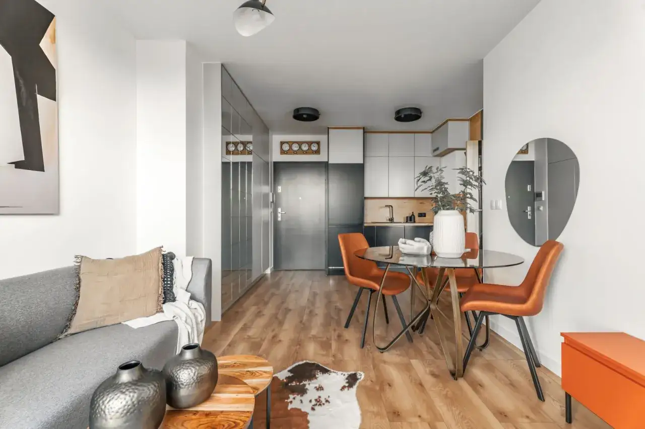Nowoczesna aranżacja mieszkania 34m2: szara sofa, okrągły st&oacute;ł z pomarańczowymi krzesłami, kuchnia i lustrzana szafa.