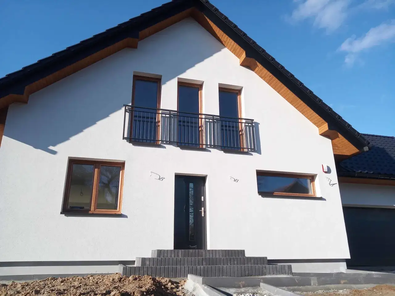 Nowy dom z białym tynkiem, antracytowym dachem i oknami w kolorze winchester. Na piętrze balkon z czarną balustradą.