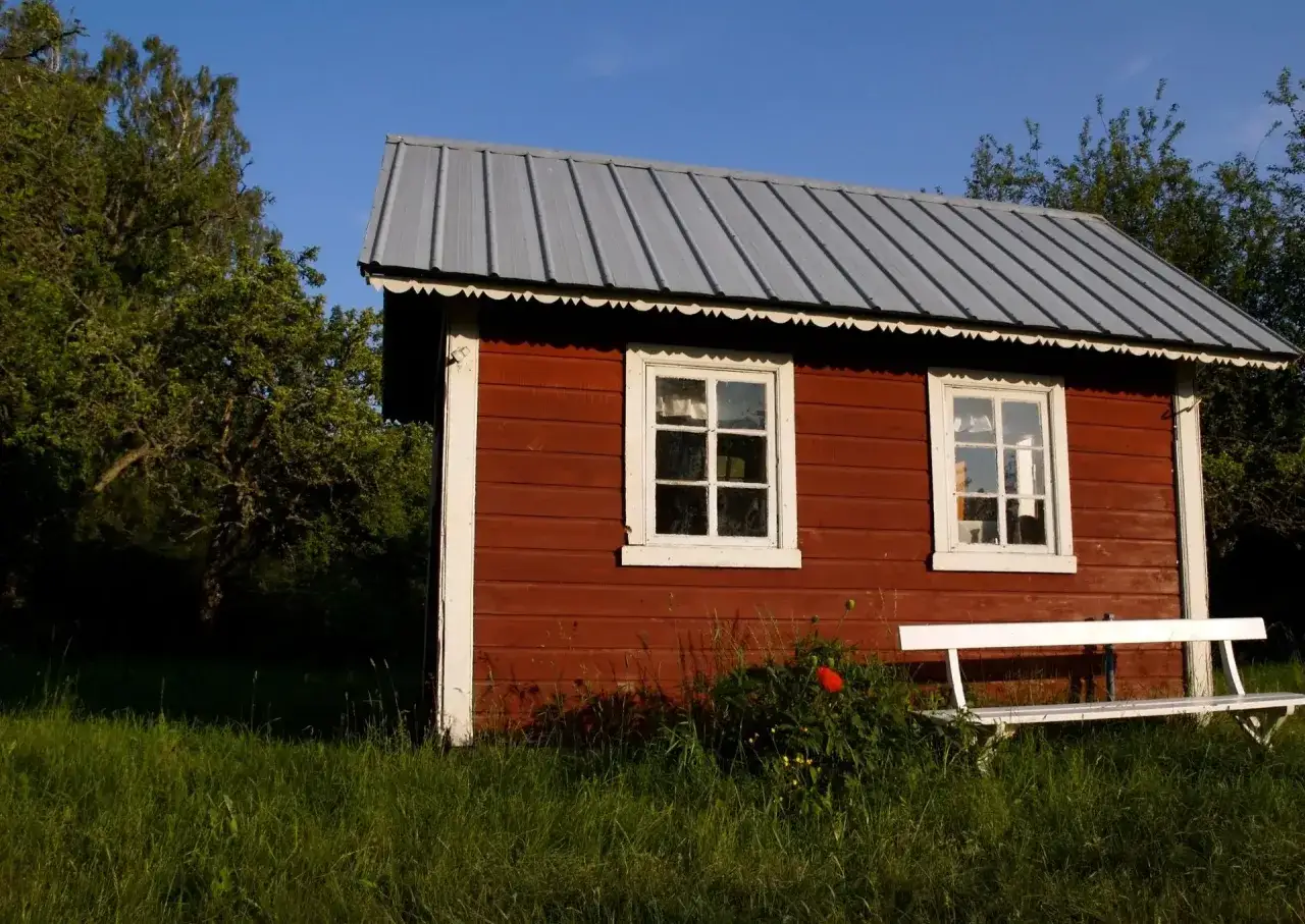 Czerwony domek letniskowy z białymi oknami i ławką. Zastanawiasz się, ile kosztuje budowa domku letniskowego drewnianego?