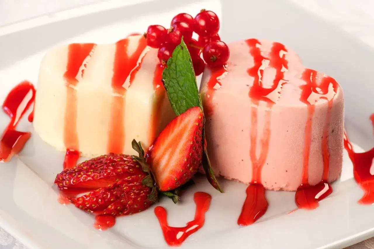 Dwa serca z panna cotta, polane sosem truskawkowym, ozdobione świeżymi truskawkami i porzeczkami.