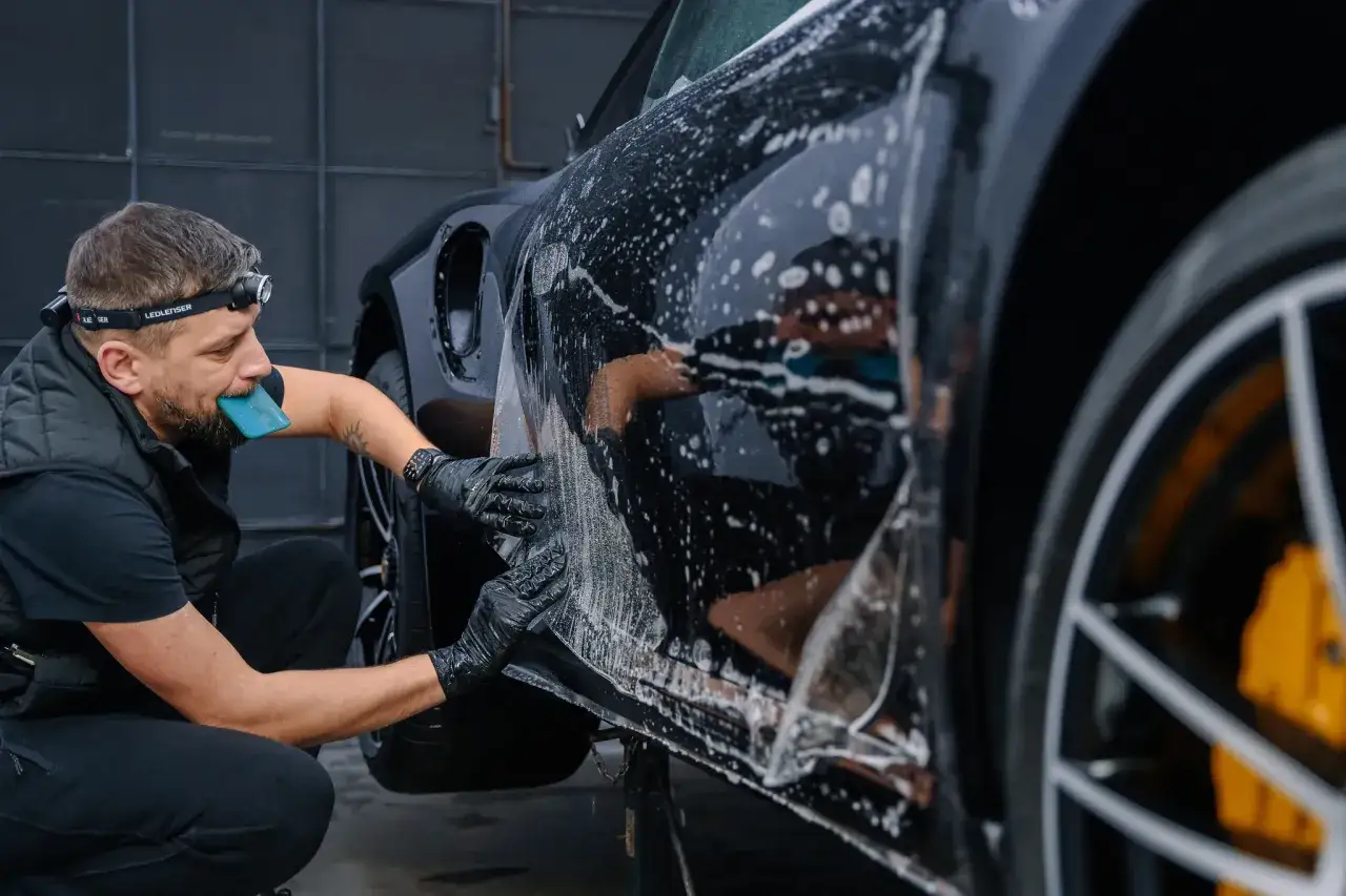 Mężczyzna w rękawiczkach i z czoł&oacute;wką na głowie aplikuje folię ochronną na błyszczący, czarny samoch&oacute;d. Detailing co to - precyzyjna ochrona lakieru.