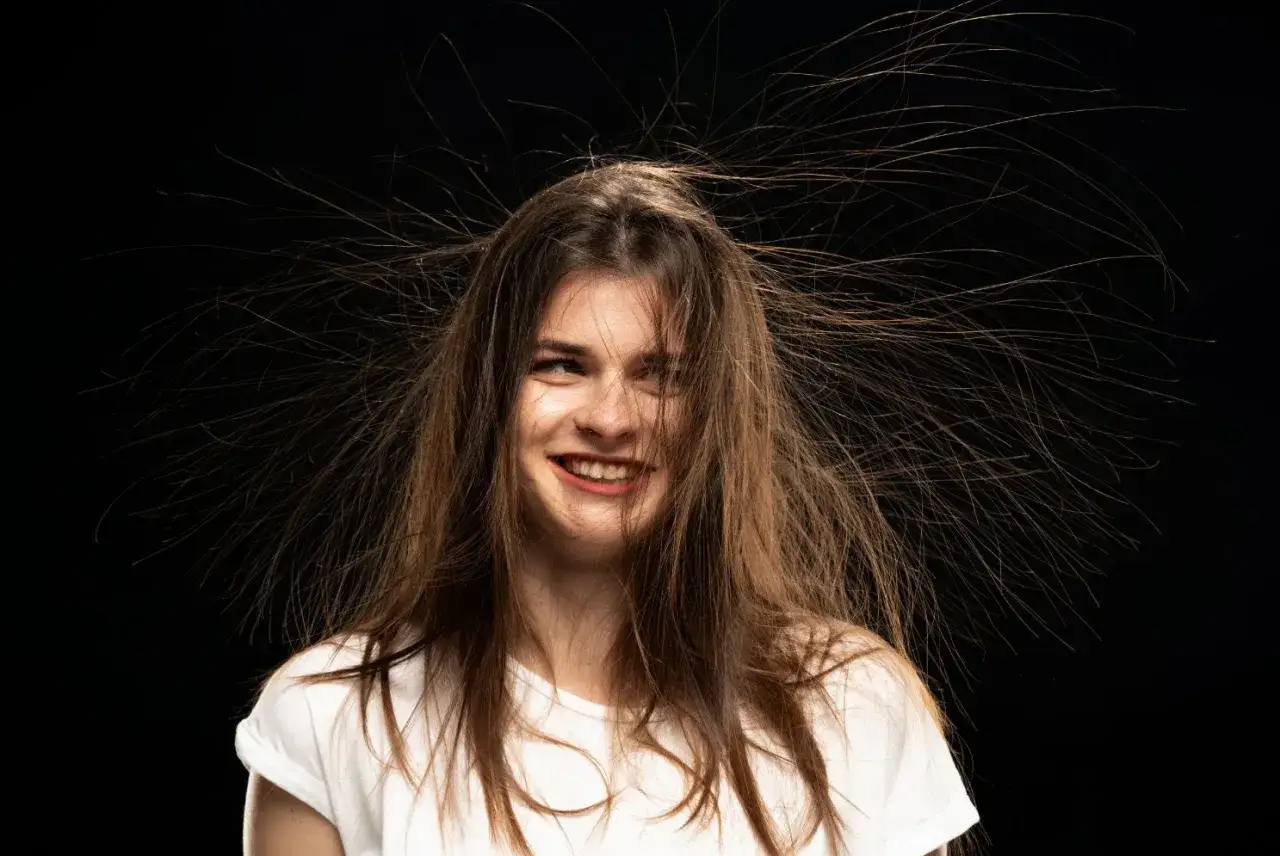 Uśmiechnięta dziewczyna z brązowymi włosami, kt&oacute;re stoją na głowie, tworząc efekt elektryzujące się włosy.