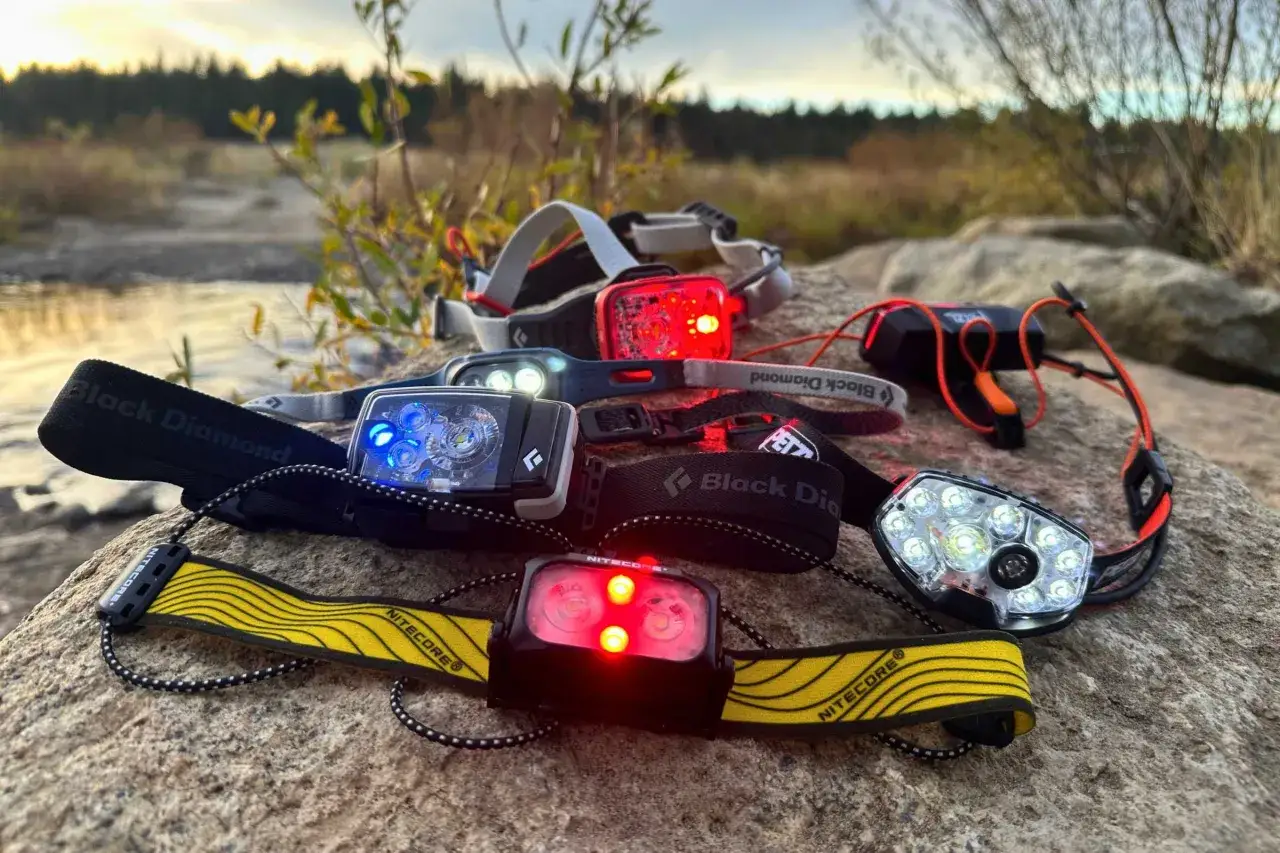 Kilka czoł&oacute;wek do biegania leży na kamieniach przy strumieniu. R&oacute;żne modele, w tym Black Diamond i Nitecore, z włączonymi światłami białymi i czerwonymi.