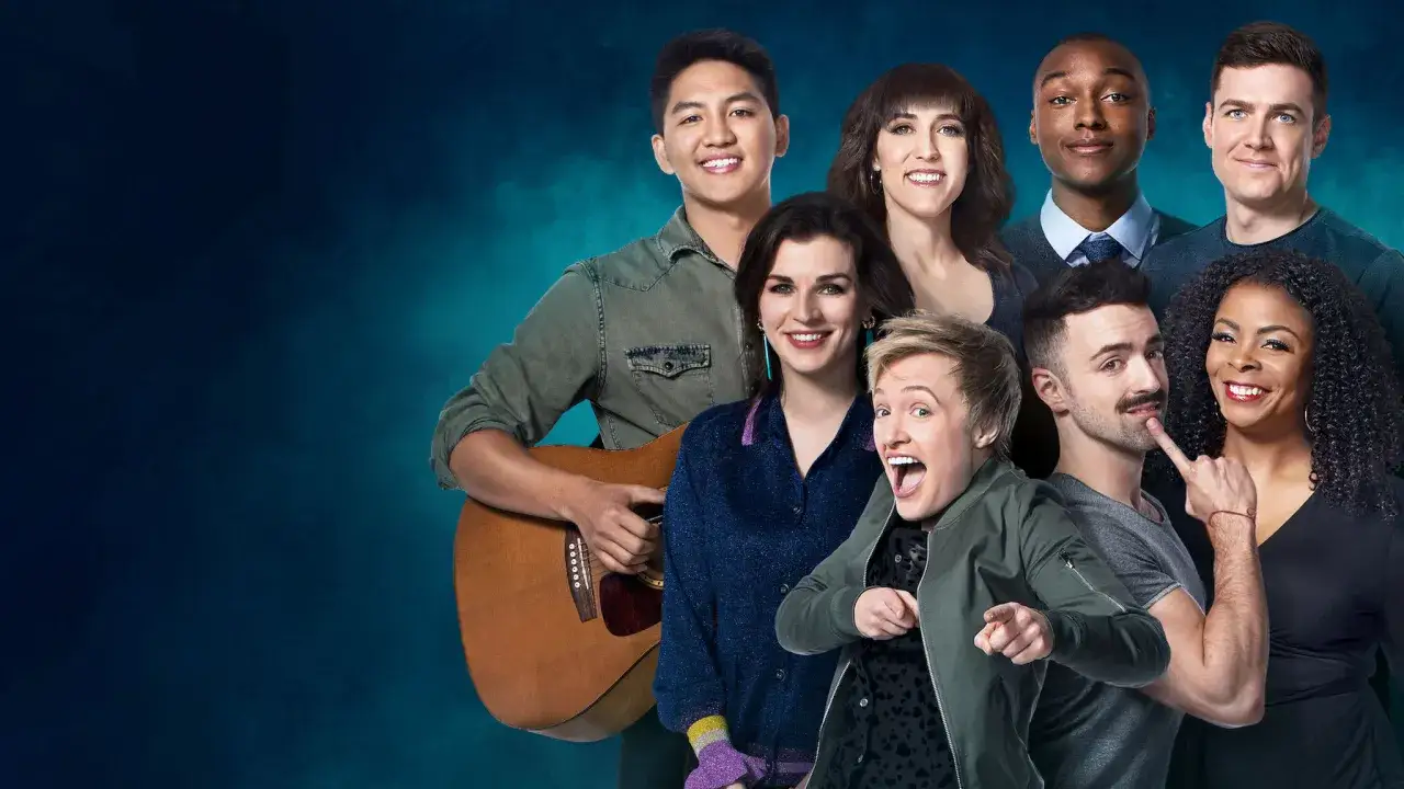 Grupa uśmiechniętych ludzi, w tym jeden z gitarą, gotowa do występu. Może to być amerykański komik stand up i jego zesp&oacute;ł.