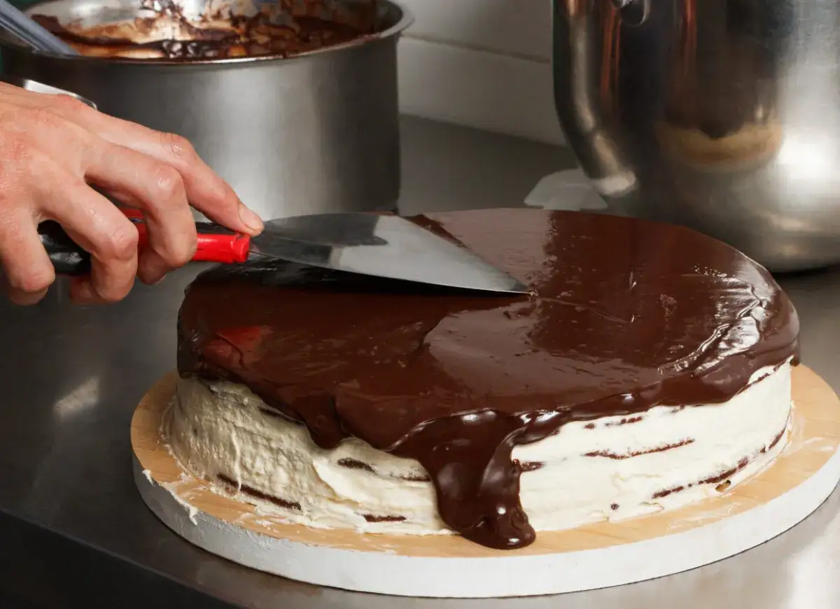 Ręka rozprowadza błyszczącą polewę czekoladową na torcie. Idealny moment, kiedy polać ciasto polewą czekoladową, by uzyskać taki efekt.