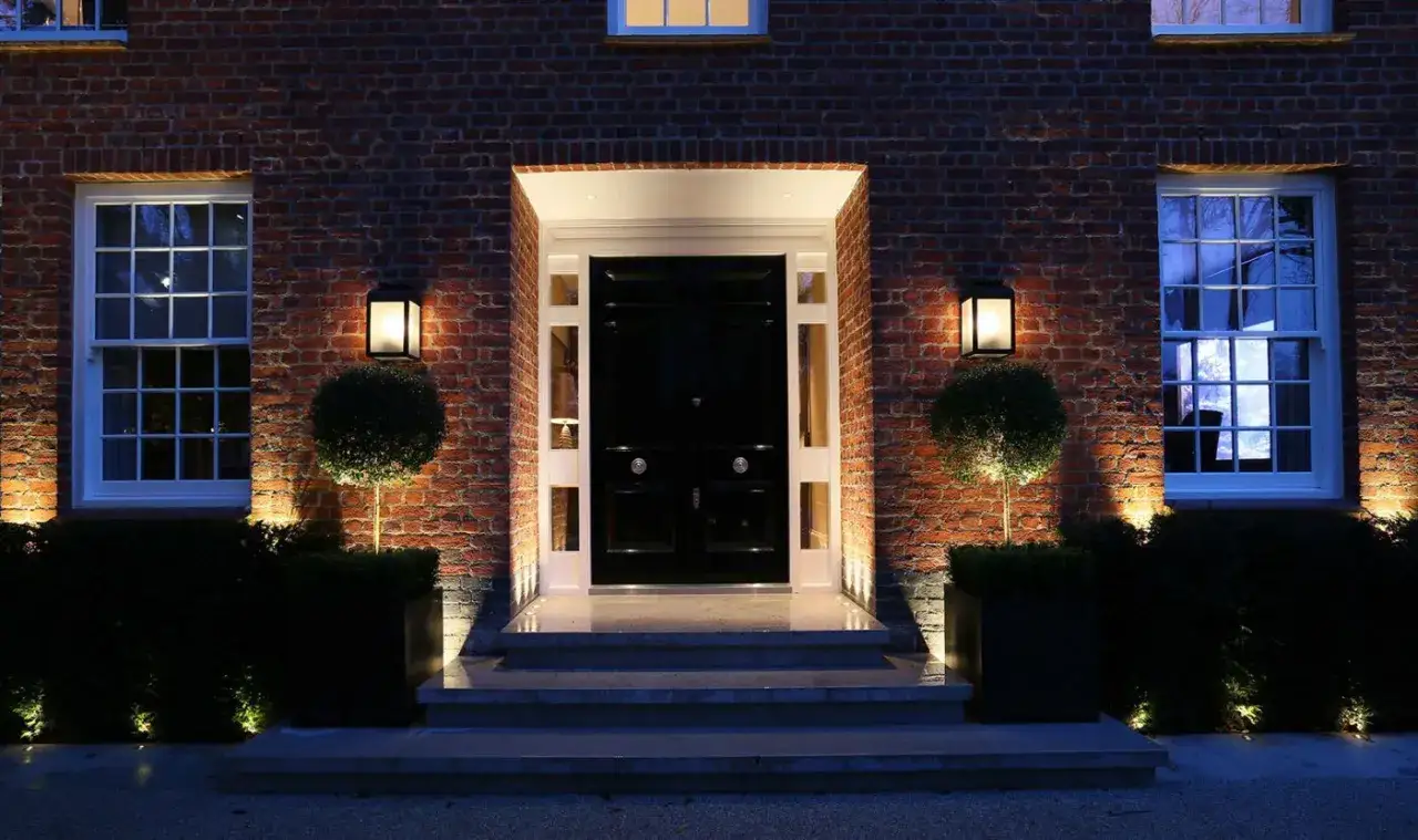 Eleganckie, punktowe jakie oświetlenie przed wejściem do domu podkreśla schody, krzewy i ozdobne drzewka, tworząc przytulną atmosferę.