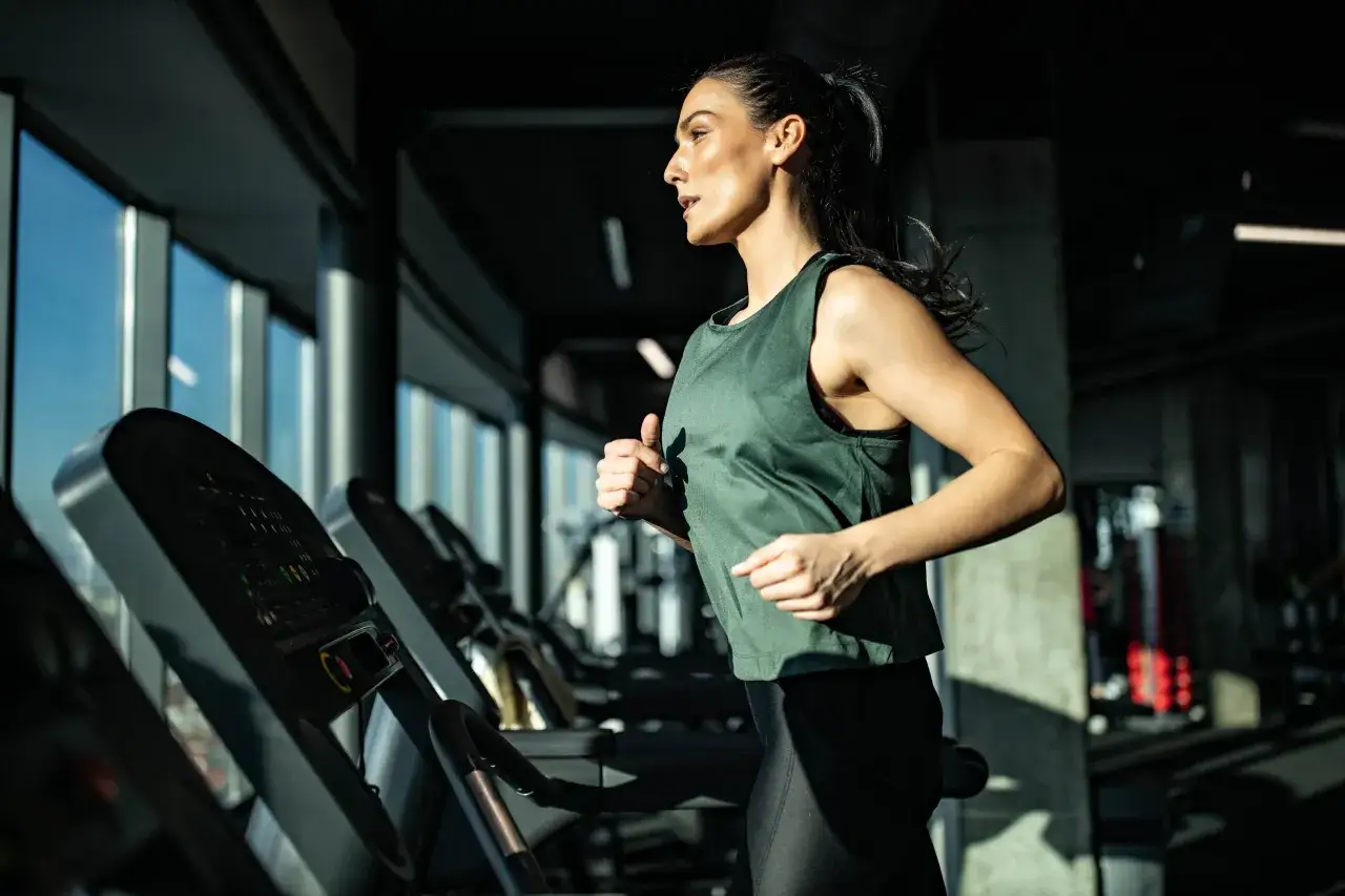 Kobieta biegnie na bieżni w siłowni. Wybór między bieżnią a orbitrekiem zależy od celów treningowych.