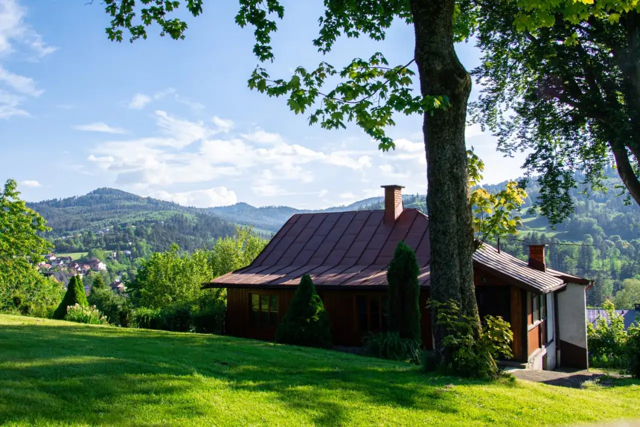 Drewniany domek z czerwonym dachem, otoczony zielenią, z widokiem na Beskid Śląski. Idealne miejsce na wypoczynek i odkrywanie beskidzkich atrakcji.