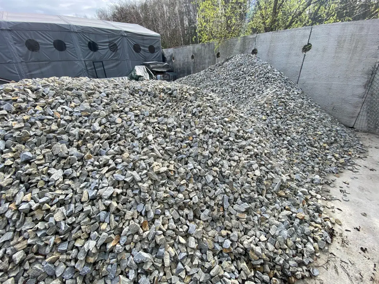 Ogromna hałda tłucznia na drodze, gotowa do budowy lub remontu. W tle szary namiot i betonowy mur.