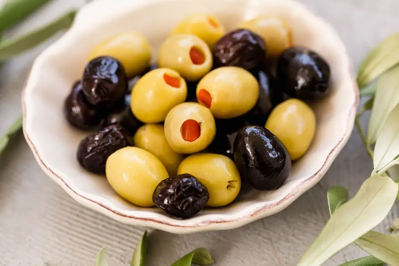 Miseczka pełna oliwek, zielonych i czarnych, z widocznymi pestkami. Ich r&oacute;żnorodne kolory oliwek zachęcają do degustacji.