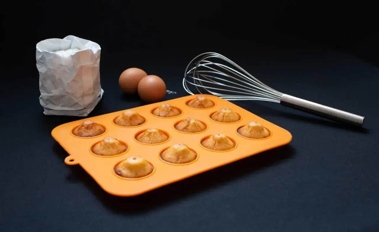Upieczone babeczki w pomarańczowej foremce silikonowej. Czy silikonowe foremki do babeczek trzeba smarować tłuszczem? Widać mąkę, jajka i trzepaczkę.