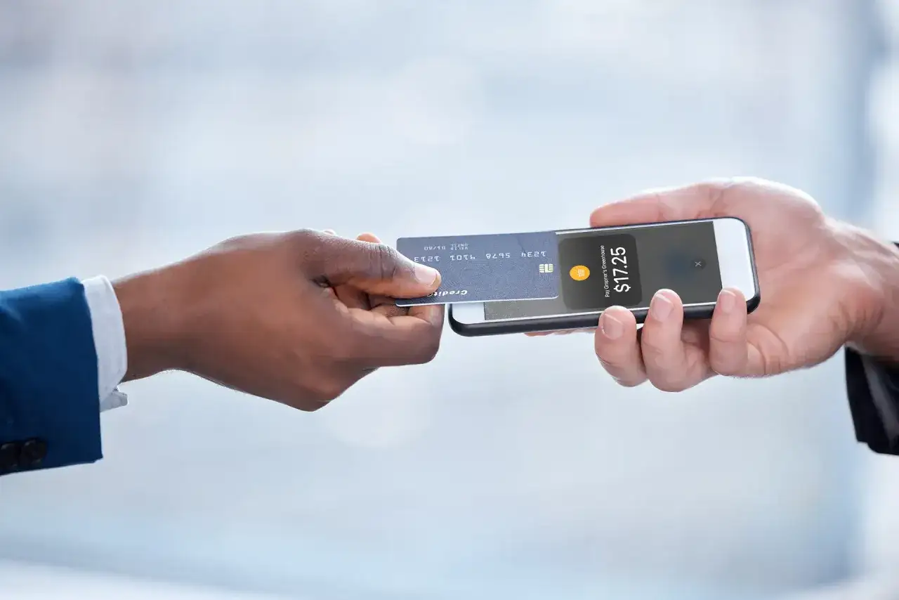 Zbliżenie na dłonie: jedna podaje kartę zbliżeniową do telefonu, na ekranie którego widnieje kwota 17.25$ i logo płatności NFC.