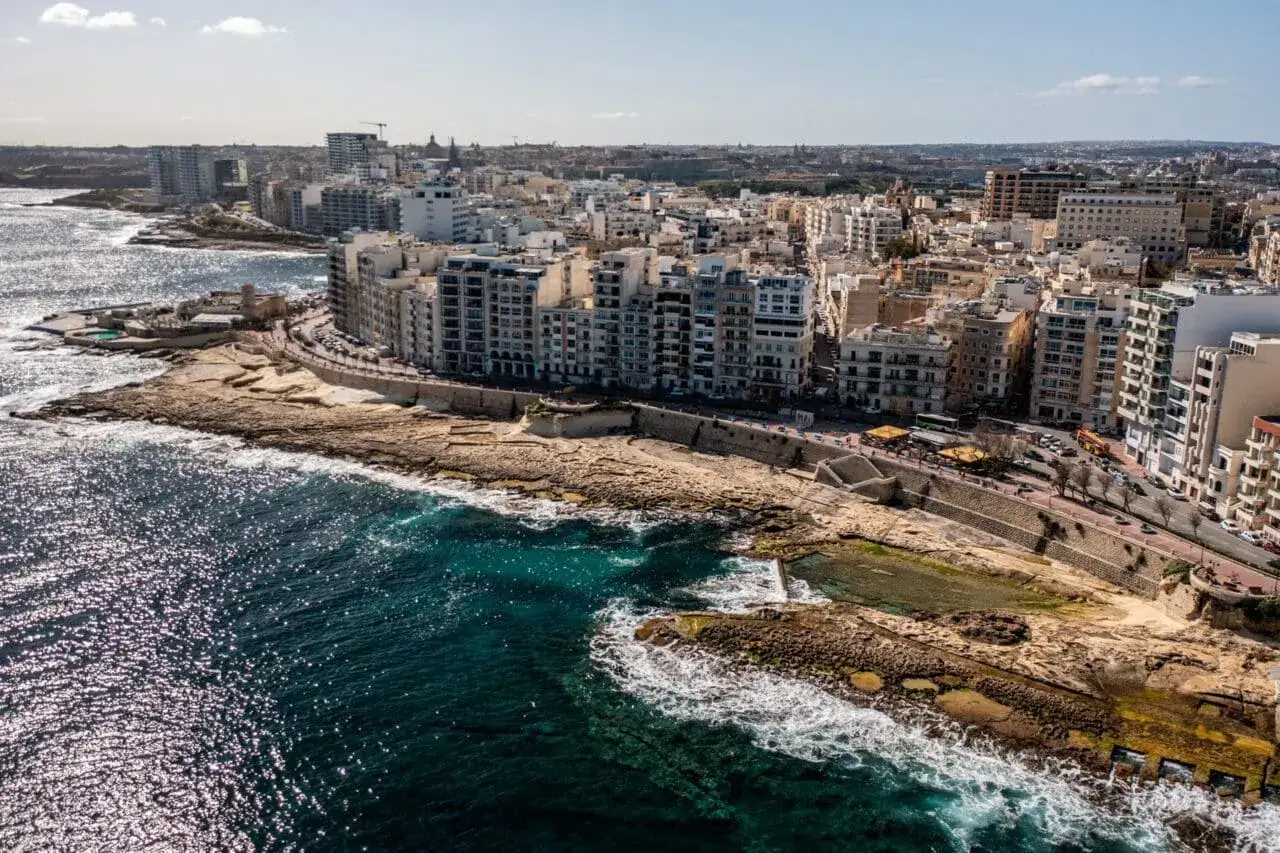 Widok na wybrzeże z budynkami mieszkalnymi i skalistym brzegiem. To miejsce, gdzie leży Malta na mapie, w otoczeniu turkusowego morza.