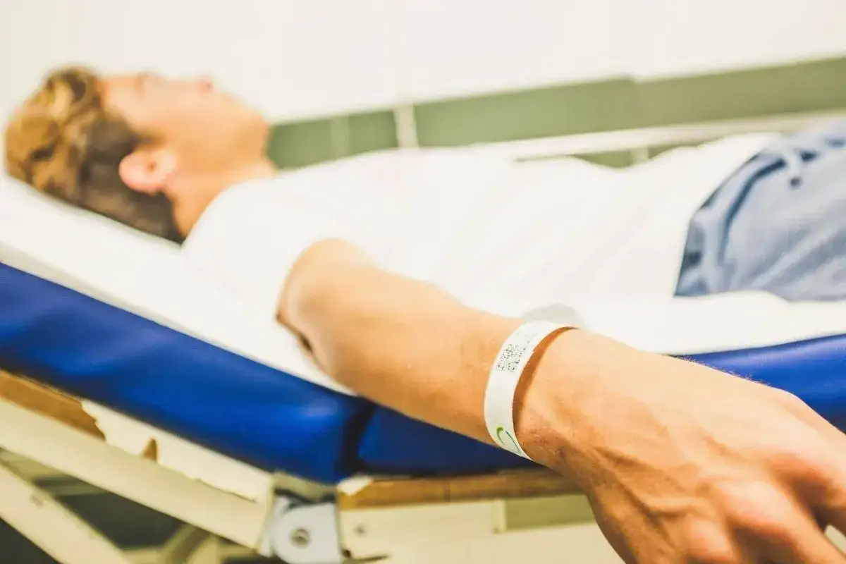 Chłopak leży na łóżku, a na jego ręce widnieje biała opaska z kodem QR. To opaska identyfikacyjna, która oznacza, co oznacza niebieska opaska na ręce w szpitalu.