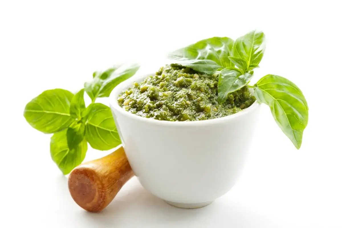 Aksamitne, zielone **pasta pesto** w białej miseczce, ozdobiona listkami bazylii. Obok moździerz.