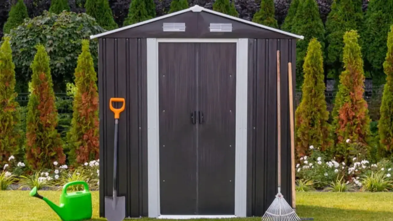 Nowy domek na narzędzia ogrodowe czeka na swoje wyposażenie. Obok stoją łopata, grabie i zielona konewka.