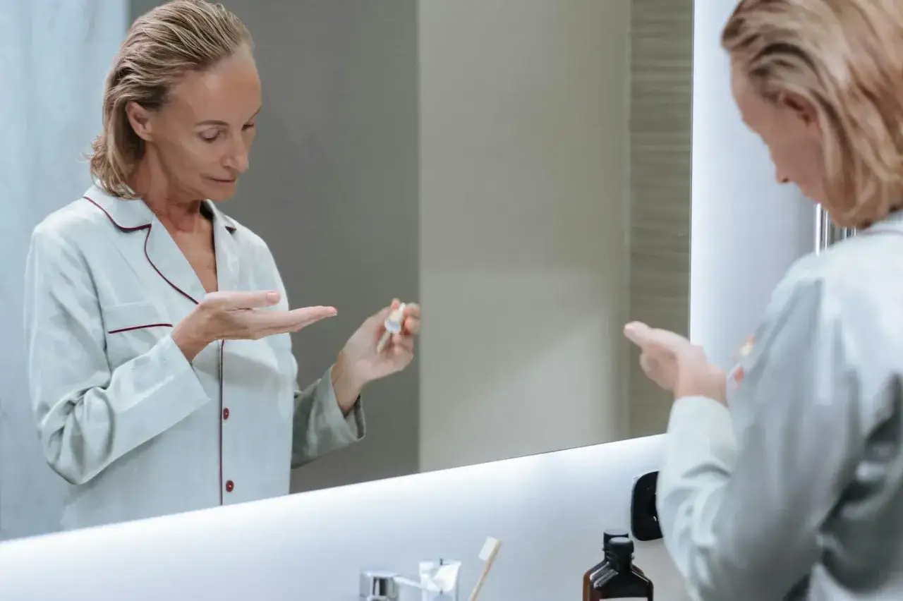 Kobieta w piżamie aplikuje serum z **retinolem na trądzik** do dłoni, patrząc w lustro.