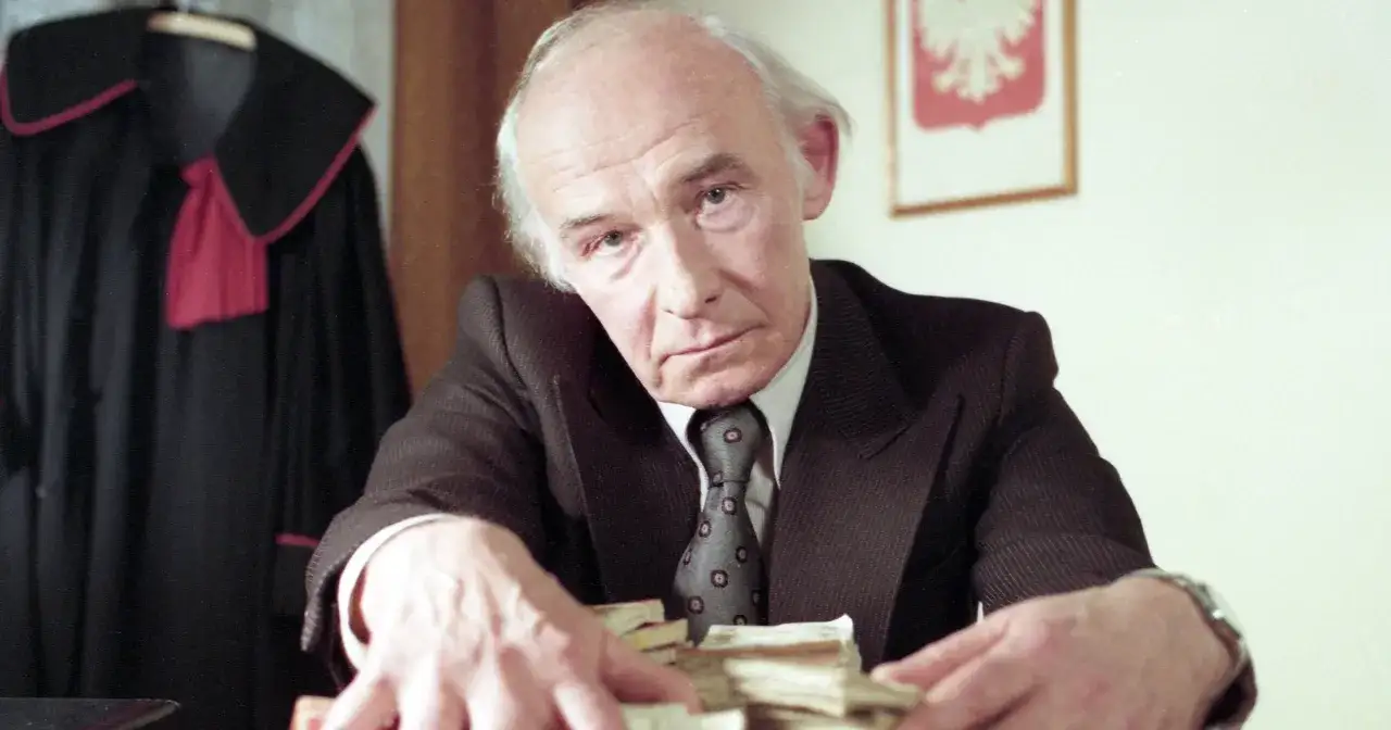 Henryk Bista w garniturze i krawacie, z rękami na stosach pieniędzy. W tle toga prawnicza.