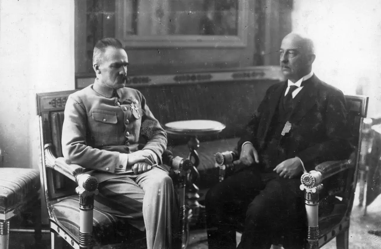 Dwóch mężczyzn siedzi w fotelach. Jeden w mundurze, drugi w garniturze. Scena z lat dwudziestych XX wieku.