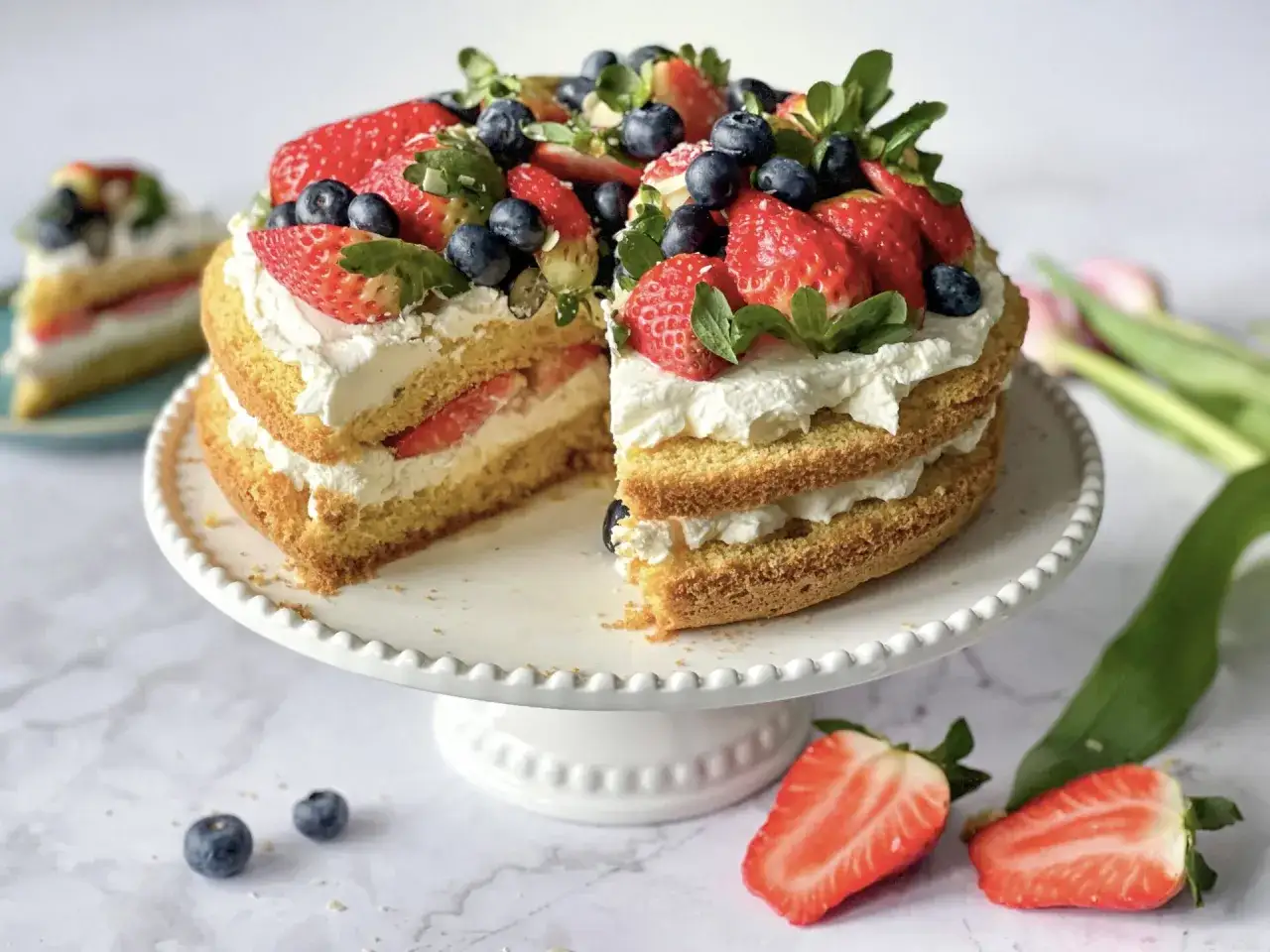 Pyszne torty bezglutenowe przepisy na lato: biszkopt przełożony kremem i świeżymi owocami, ozdobiony truskawkami i borówkami.