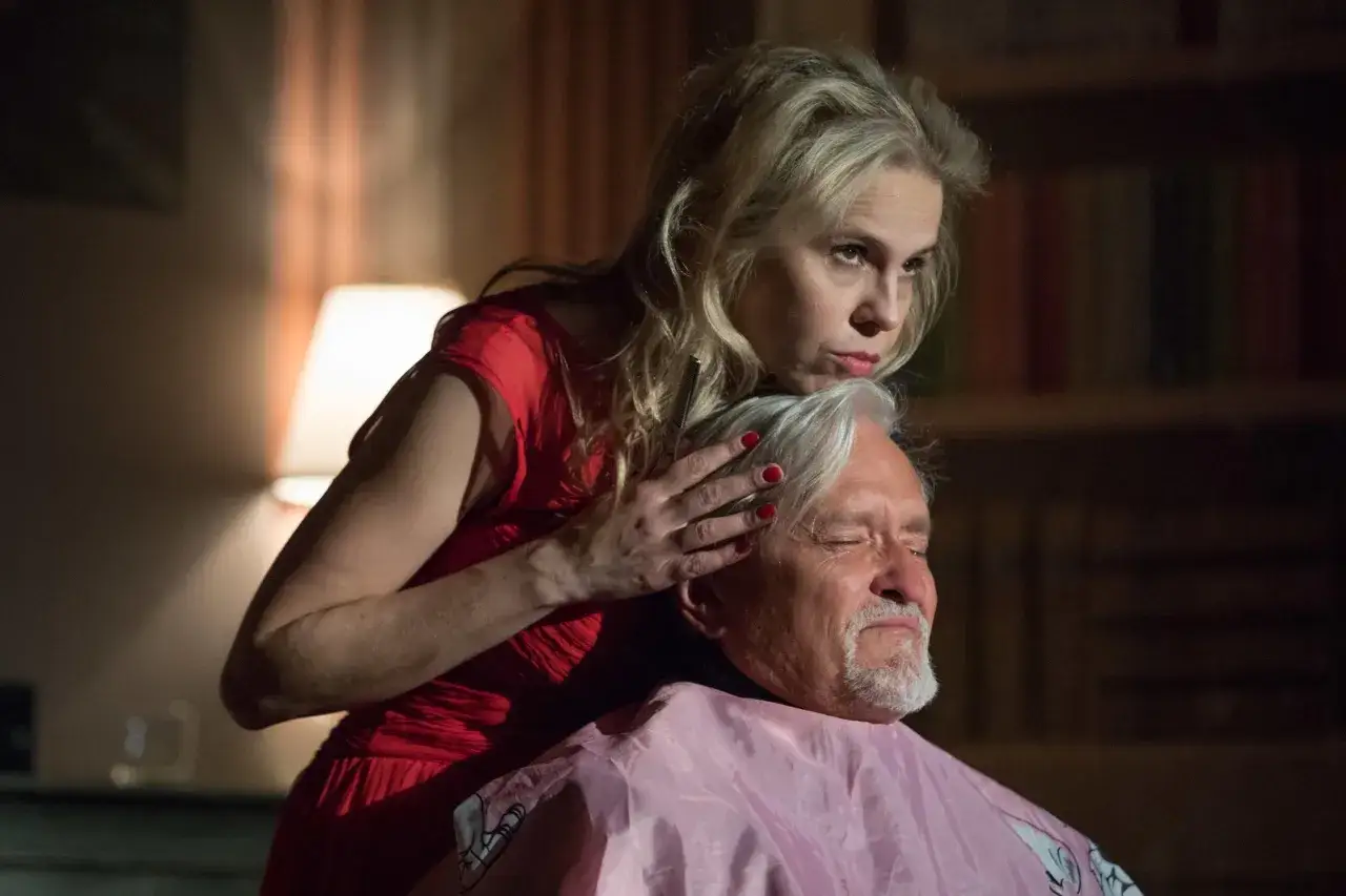 Kobieta w czerwonej sukience czesze siwowłosego mężczyznę. To scena z filmu, gdzie trwa edukacja Rity.