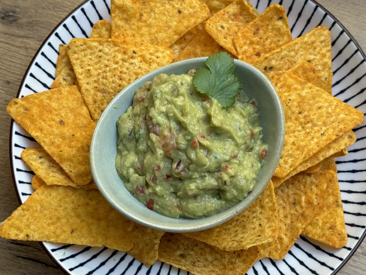 Pyszne guacamole jak jeść? Nachosami! Kremowy dip z awokado, pomidorów i kolendry, podany z chrupiącymi chipsami kukurydzianymi na talerzu w paski.