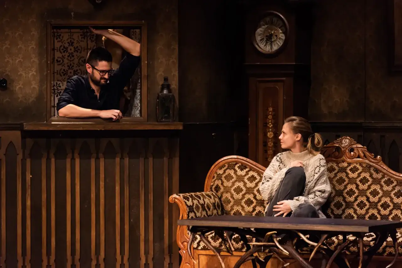 Scena z Teatru Kwadrat w Warszawie. Mężczyzna w okularach wygląda z okienka, kobieta siedzi na kanapie. Sprawdź repertuar!