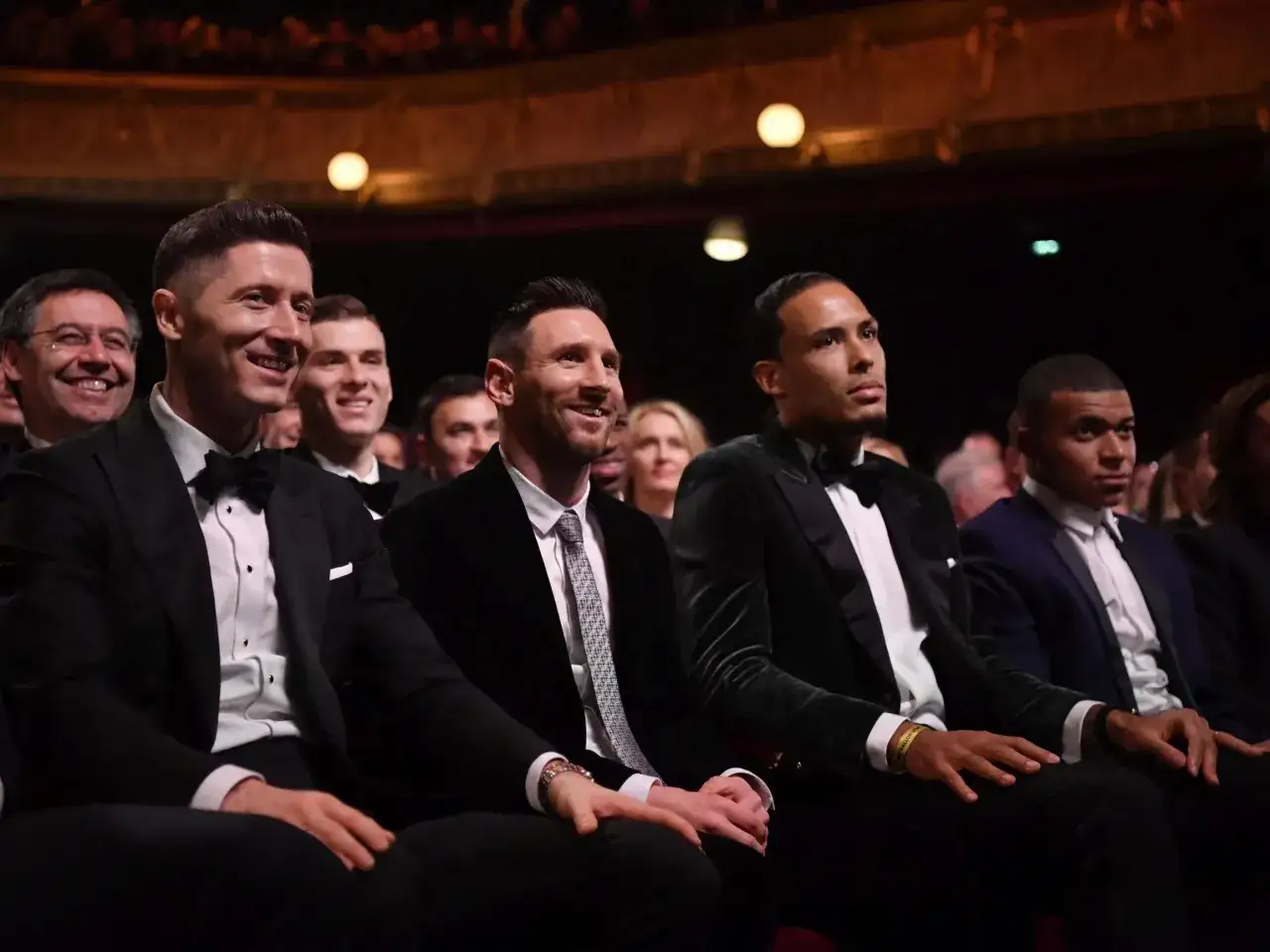 Piłkarze w eleganckich garniturach, w tym Lewandowski, Messi, Van Dijk i Mbappé, siedzą na gali Złotej Piłki, czekając na ogłoszenie wyników.