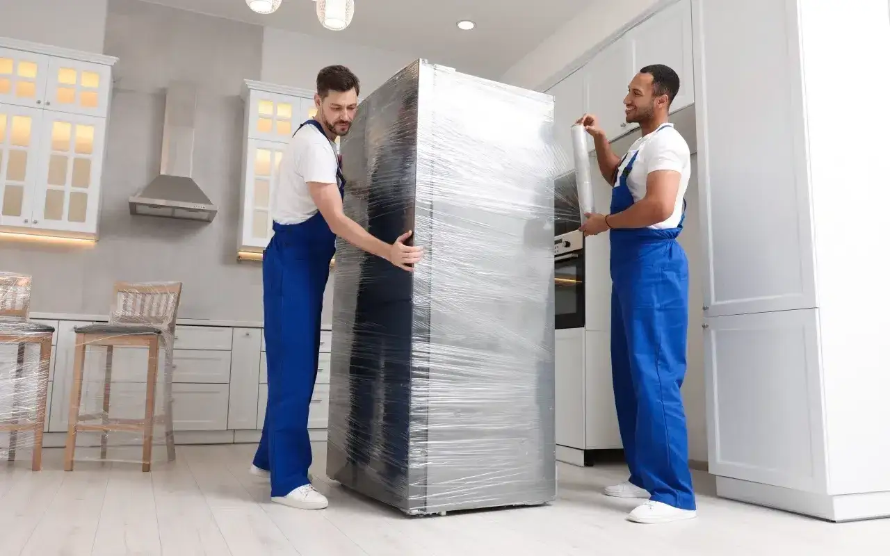 Dw&oacute;ch mężczyzn w niebieskich kombinezonach zabezpiecza lod&oacute;wkę folią, pokazując, jak przewozić lod&oacute;wkę bezpiecznie.