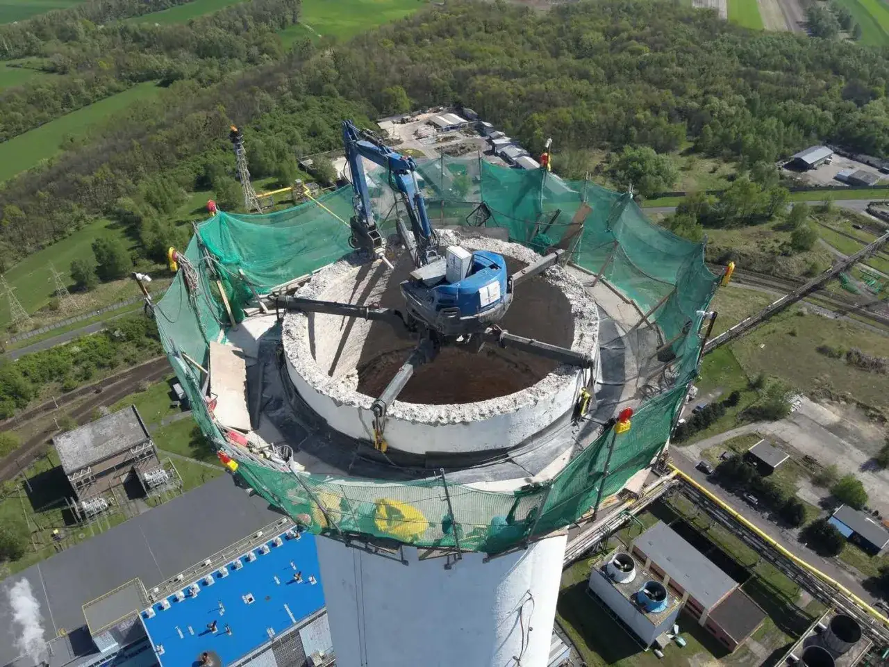 Maszyna rozbiórkowa pracuje na szczycie komina. Zastanawiasz się, ile kosztuje rozbiórka komina i postawienie nowego?