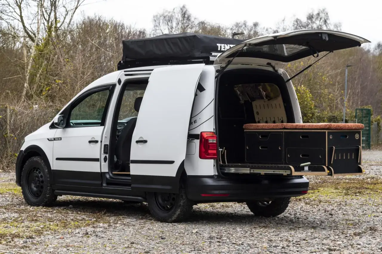 Biały caddy kamper z otwartymi drzwiami i bagażnikiem, gotowy na przygodę. Na dachu namiot dachowy.