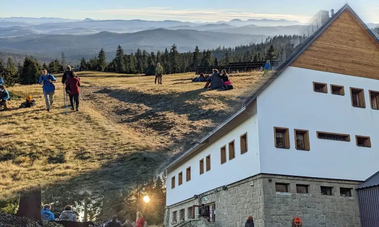 Grupa turystów na szlaku w górach, odpoczywają przy schronisku. W tle rozległe pasma górskie.
