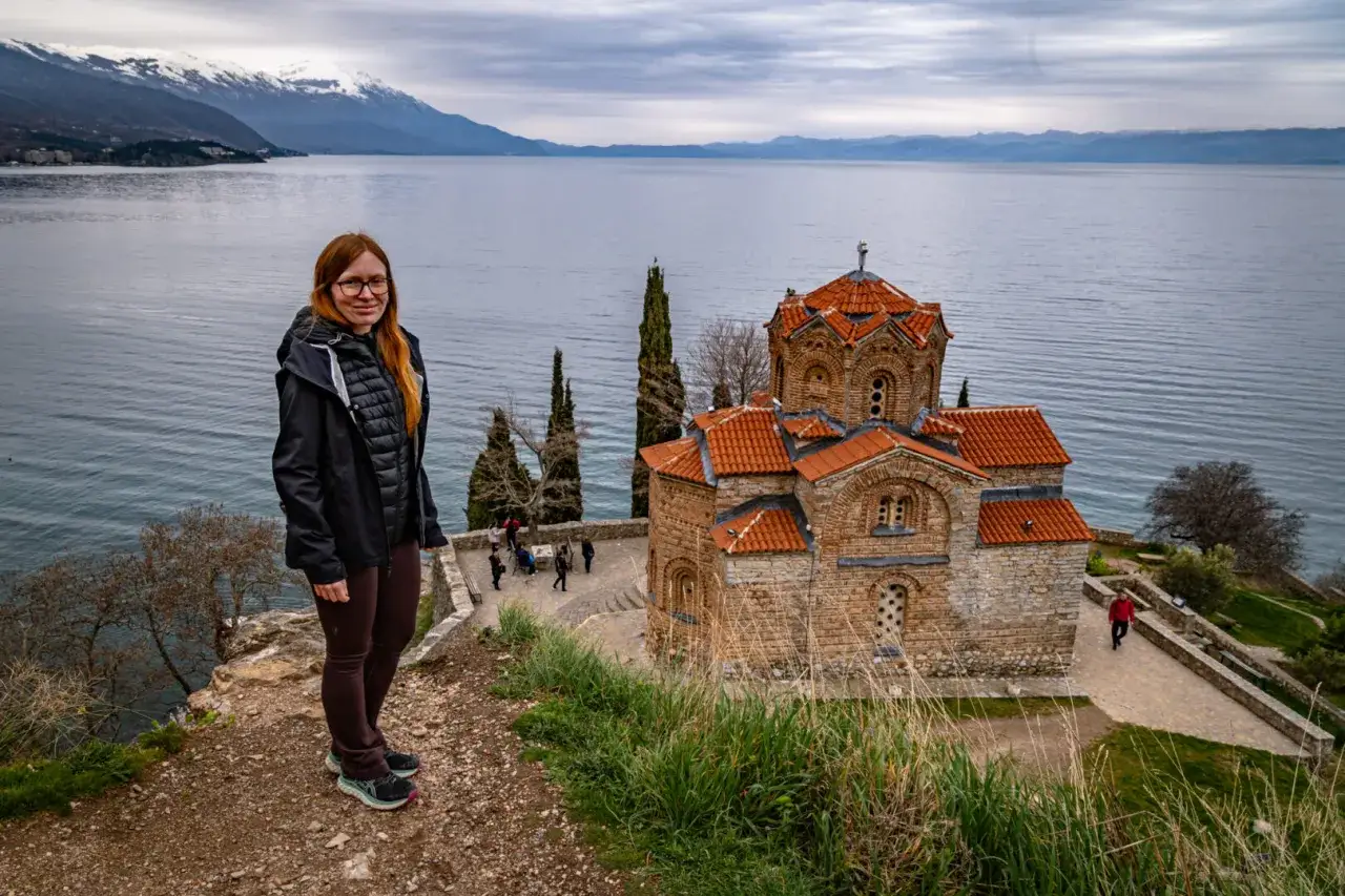 Kobieta z rudymi włosami pozuje na tle malowniczego jeziora Ochryd. W oddali widać ośnieżone g&oacute;ry i zabytkową cerkiew.
