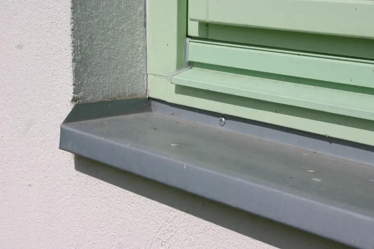 Szary parapet zewnętrzny zamontowany pod zielonym oknem. Detale pokazują, jak prawidłowo zamontować parapet zewnętrzny, z widocznym wkrętem.