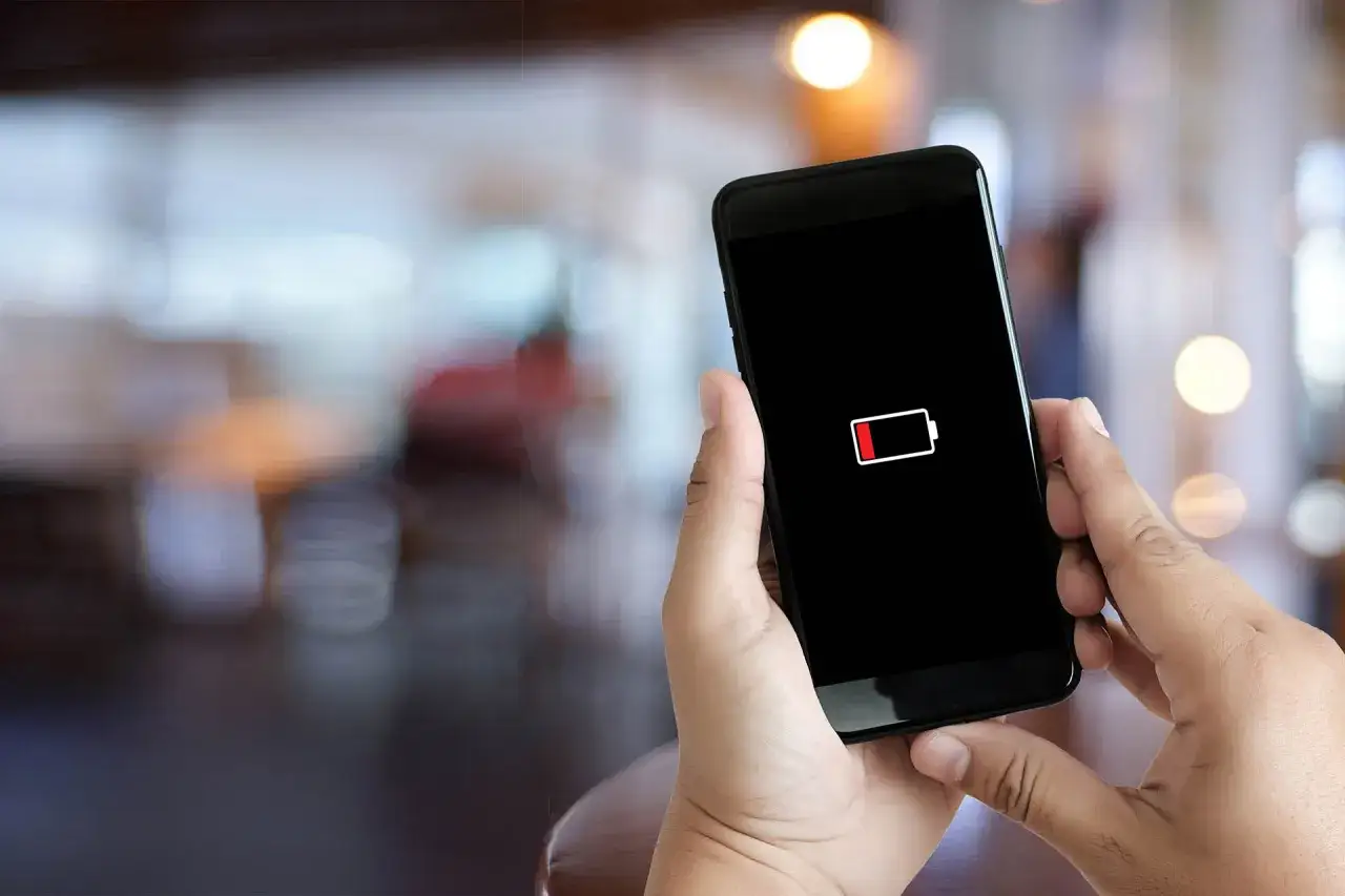 Dłoń trzyma smartfon z ikoną niskiego poziomu baterii. Aby oszczędzić energię, warto wiedzieć, jak wyłączyć aplikacje w tle.