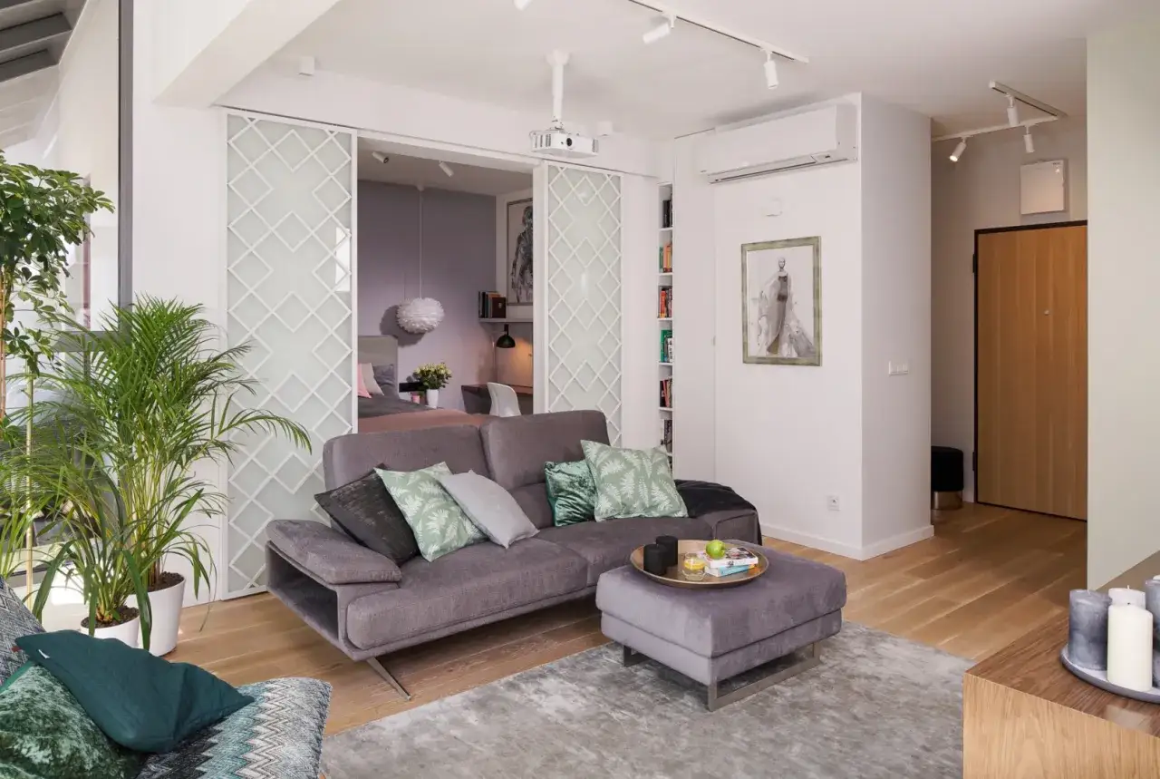 Nowoczesna aranżacja mieszkania 50m2 w bloku. Szara kanapa, ozdobne drzwi przesuwne, rośliny i stylowe dodatki tworzą przytulną przestrzeń.