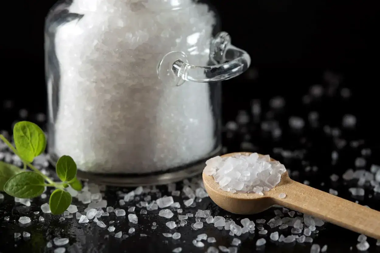 Drewniana łyżka pełna kryształków soli, obok szklanego naczynia z solą. Zastanawiasz się, ile waży łyżka soli?