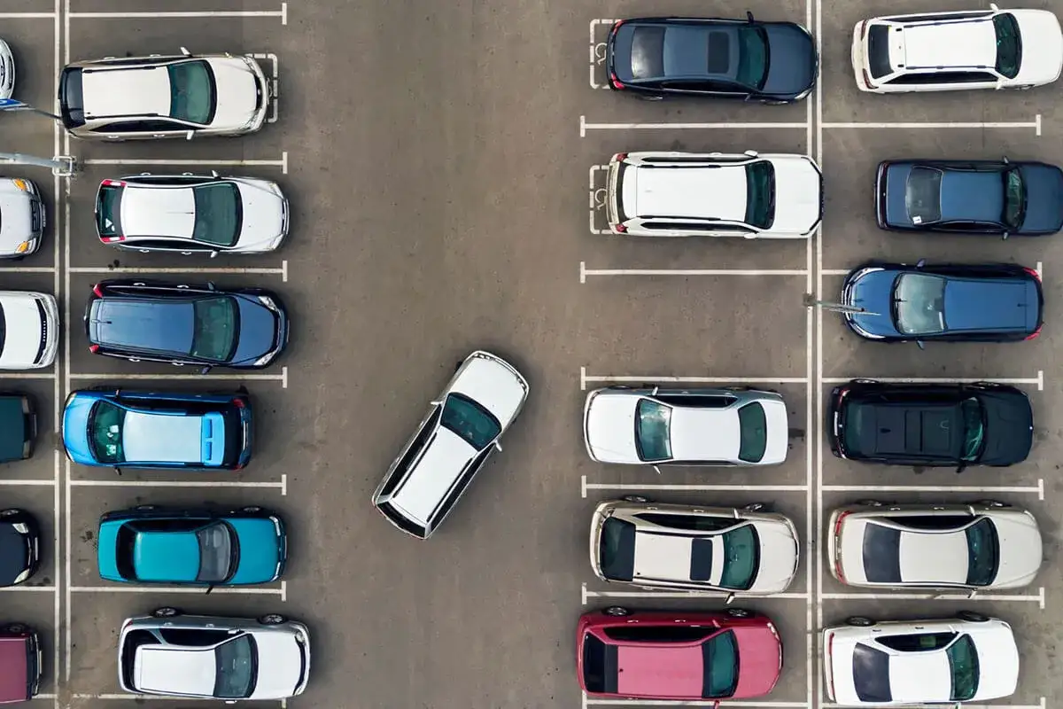 Widok z góry na parking. Samochody zaparkowane prostopadle w rzędach. Jeden biały samochód stoi pod kątem.