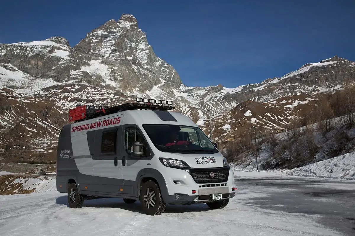 Ducato 4x4 Expedition gotowy na podb&oacute;j g&oacute;rskich szlak&oacute;w. Biało-szary kamper z bagażnikiem dachowym i dodatkowym oświetleniem na tle ośnieżonych szczyt&oacute;w.