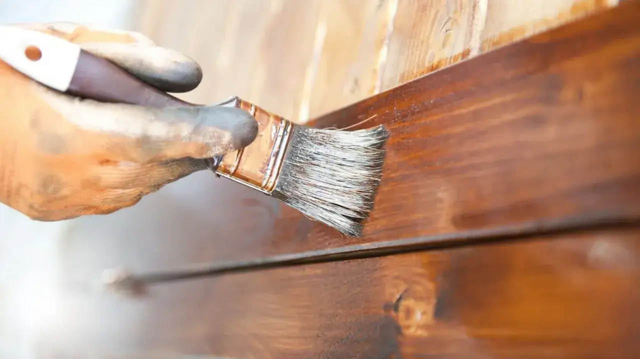 Ręka w rękawicy nakłada ciemnobrązową lakierobejcę na drewno. Czas schnięcia lakierobejcy zależy od wielu czynników.