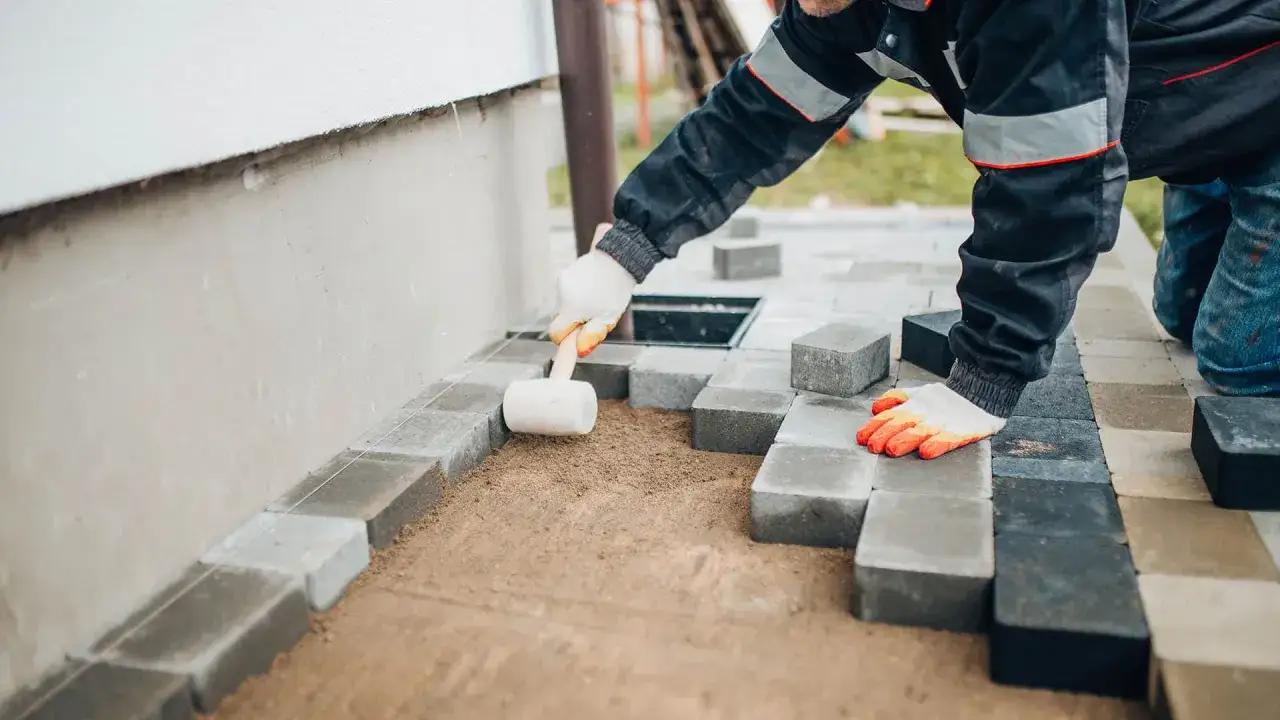 Ręce w rękawiczkach układają kostkę brukową, tworząc opaskę z kostki brukowej przy ścianie domu.