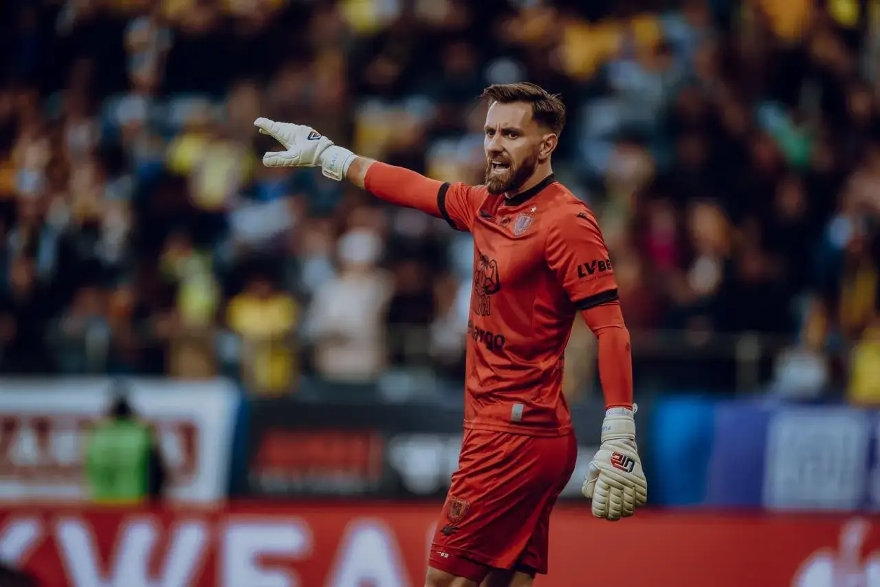 Bramkarz Piasta Gliwice w czerwonym stroju, w rękawicach, wskazuje kierunek. To jeden z kluczowych zawodników Piasta Gliwice.