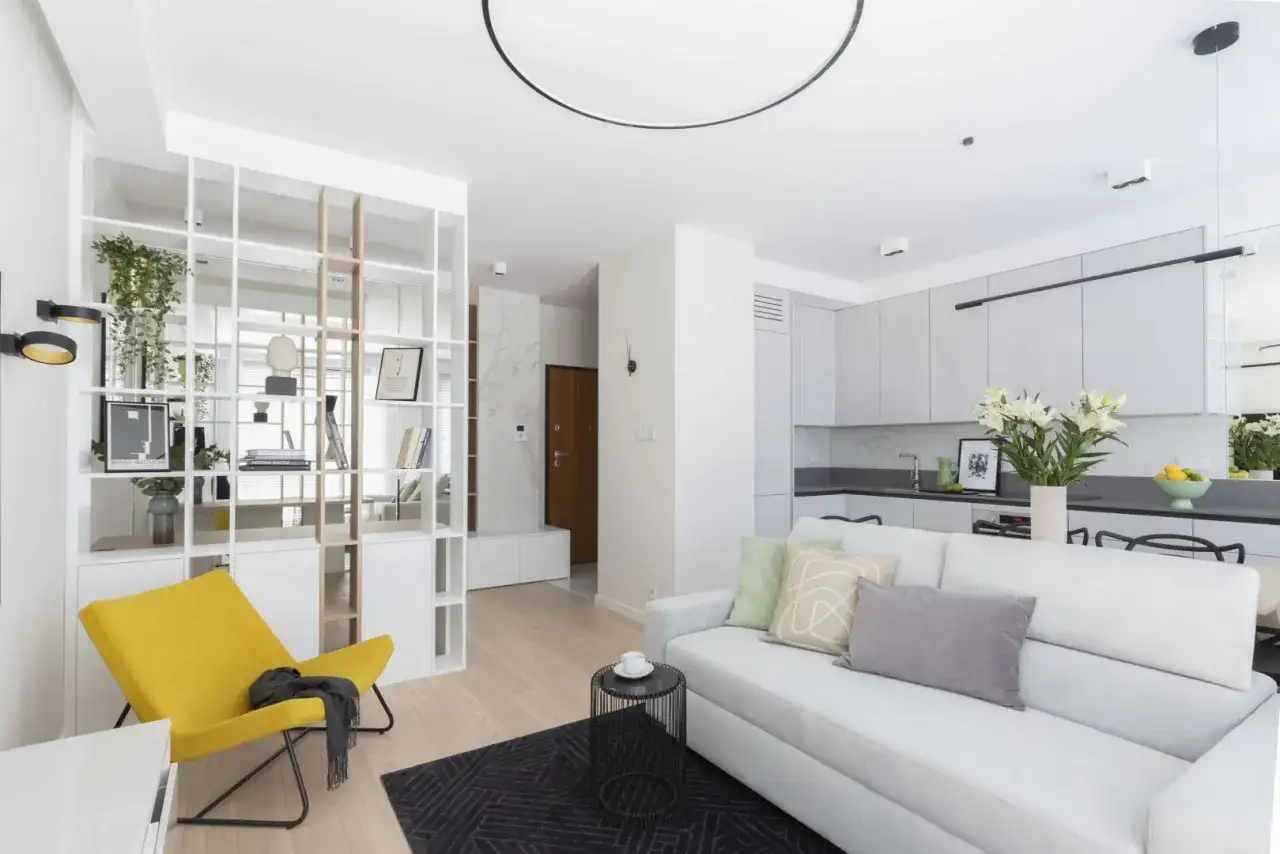 Nowoczesne oświetlenie małego pokoju dziennego z kuchnią w tle. Jasna sofa, żółty fotel i regał dzielący przestrzeń.