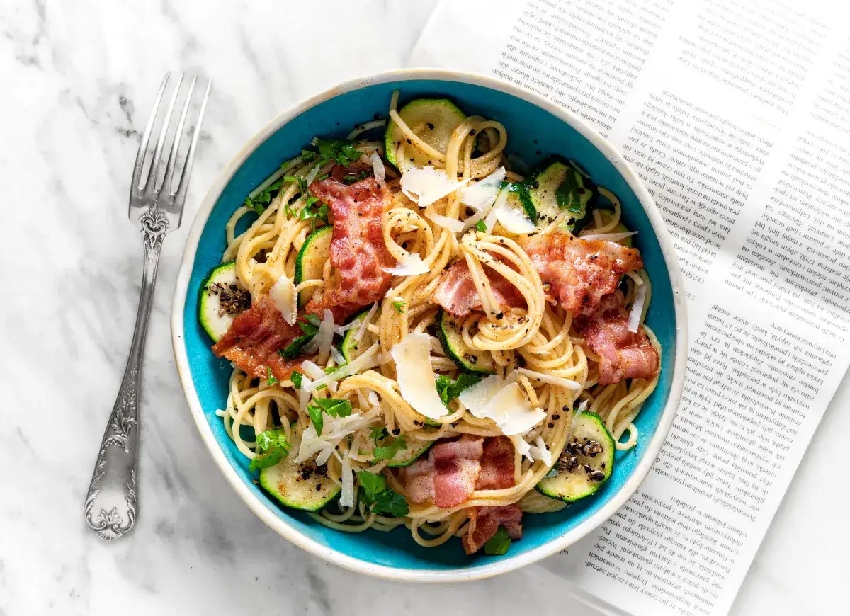 Pyszna carbonara z cukinią, podana w niebieskiej misce, z chrupiącym boczkiem i parmezanem. Obok leży widelec i otwarta książka.