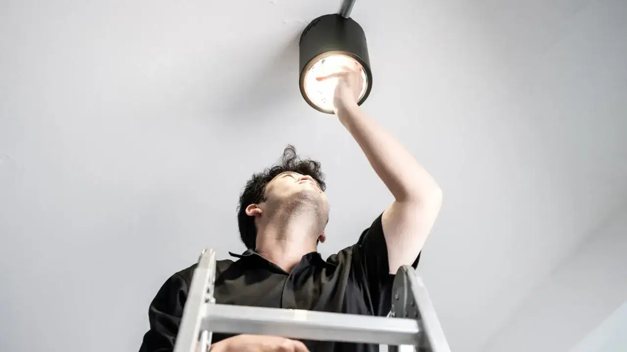 Mężczyzna na drabinie wymienia żarówkę led w oprawie sufitowej.