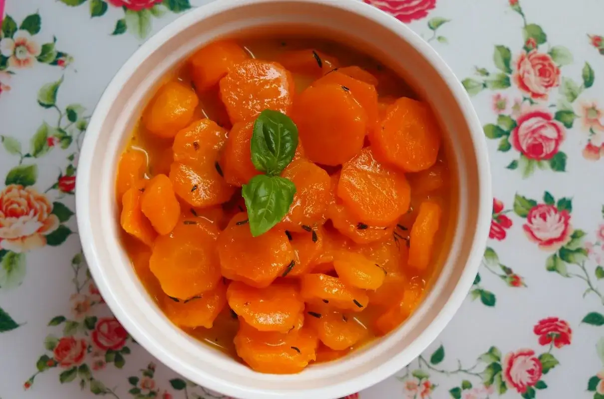 Ciepła, duszona marchewka do obiadu, udekorowana listkiem bazylii, podana w białej miseczce na kwiecistym obrusie.