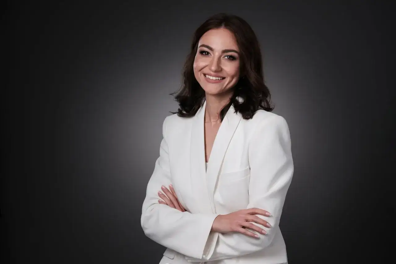 Uśmiechnięta kobieta w białej marynarce, z rękami skrzyżowanymi na piersi. To profesjonalna fotografia ludzi.